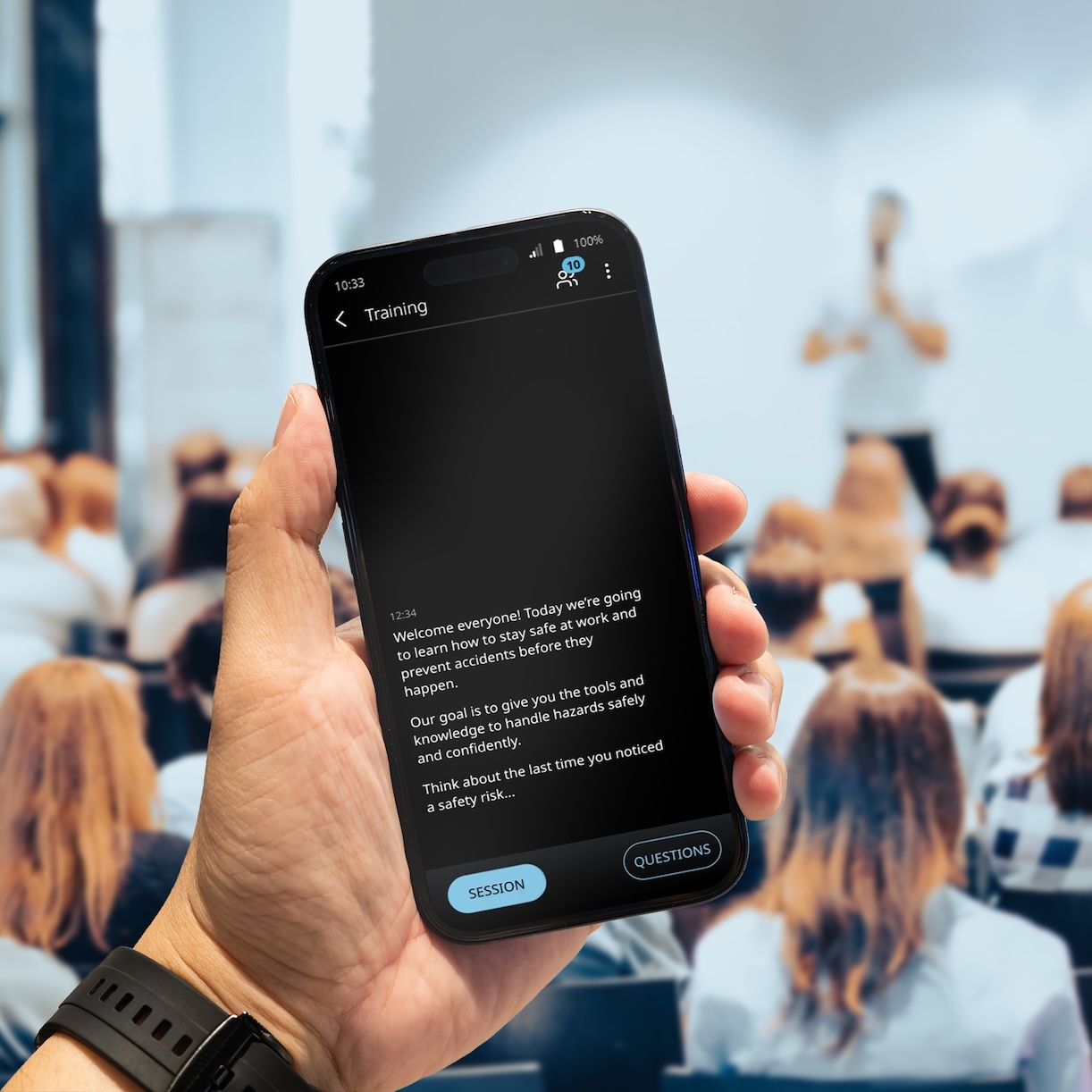 En una sala llena de gente se está impartiendo una conferencia. Un hombre sostiene en la mano un teléfono en el que se muestra la traducción mediante Vasco Audience.