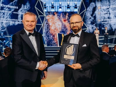 Un plano medio-largo de dos hombres vestidos de gala que se dan la mano. El hombre de la derecha, Maciej Góralski, sostiene un premio enmarcado con un emblema dorado, que identifica como el premio al «Empresario del Año 2022». En el fondo, difuminado, se ve un escenario con estructuras geométricas abstractas y motivos gráficos dinámicos en tonos azules y rojo anaranjado. Varios invitados están sentados en las mesas del salón de actos, que está en penumbra.