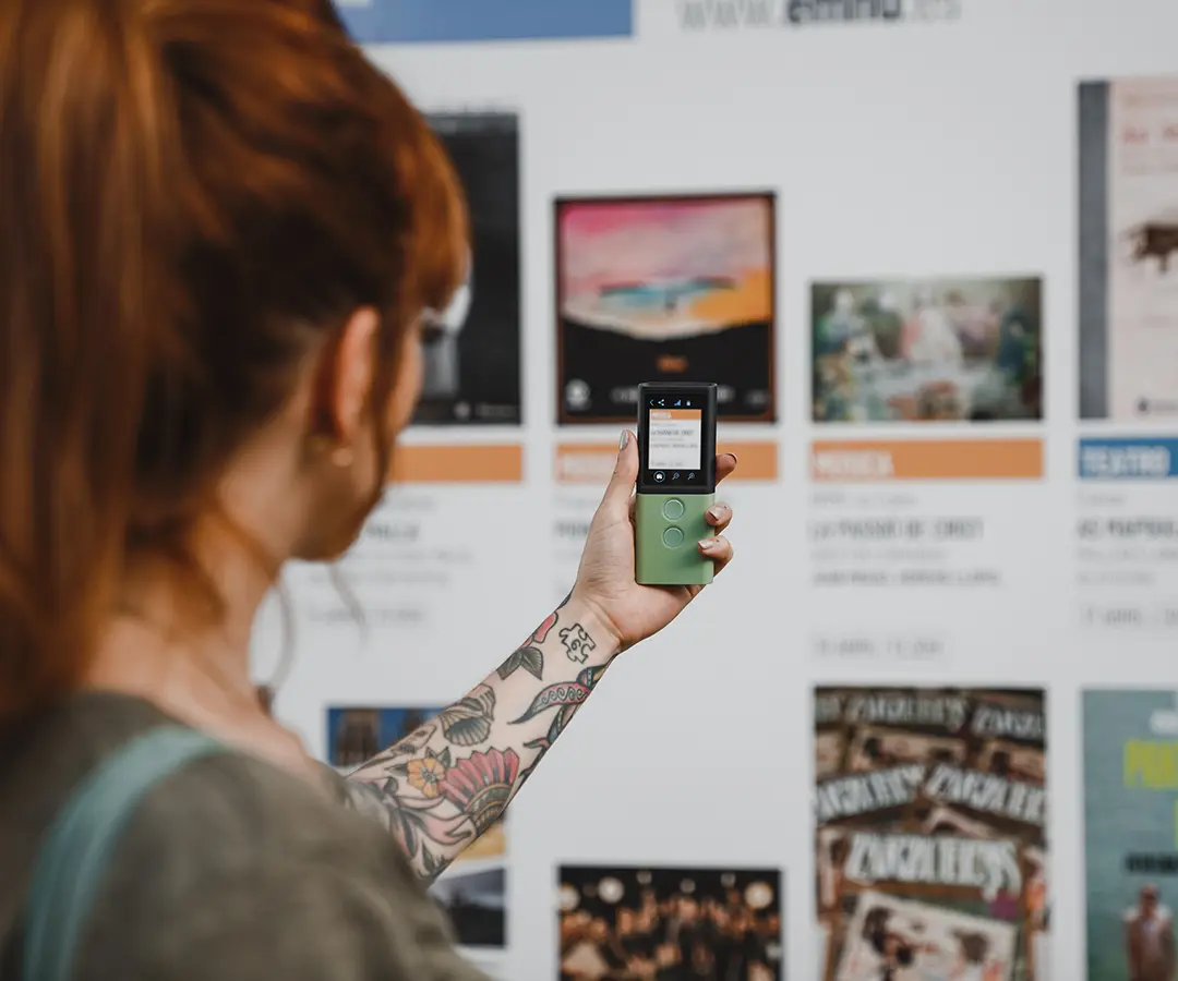 Una persona pelirroja con un tatuaje floral en el brazo sostiene un dispositivo digital verde, apuntando hacia un tablón con varios carteles e información sobre el evento.