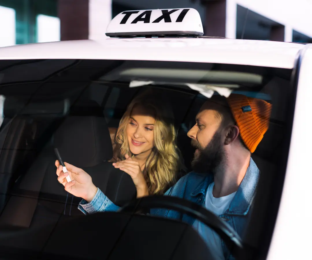 Un hombre se sienta en el asiento del conductor de un taxi y muestra algo a una mujer del asiento trasero en un traductor que lleva en la mano. La mujer sonríe con interés. En el techo del coche se ve una señal de taxi.