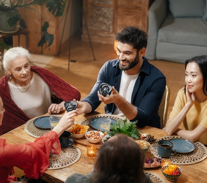 Un grupo de personas de distintos países se reunía alrededor de una mesa, disfrutando de una comida y participando en animadas conversaciones, creando un ambiente cálido y acogedor mientras se pasaban unos a otros los auriculares Traductor E1.