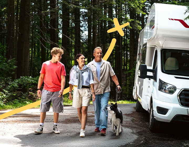 Una familia de tres —dos padres y un niño— con un perro, se alejan de su autocaravana aparcada en un día soleado. Sonríen, charlan y disfrutan de la compañía mutua.