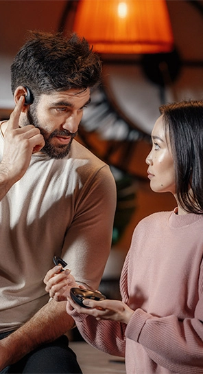 Un hombre que lleva puesto el auricular Traductor E1, está de pie junto a una mujer que sostiene el dispositivo E1, mostrando sus potentes altavoces y su micrófono inteligente.