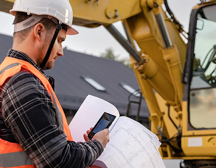 Un hombre con casco y ropa de trabajo que está tomando una foto de los planos de construcción con el Traductor V4, con una excavadora visible al fondo.