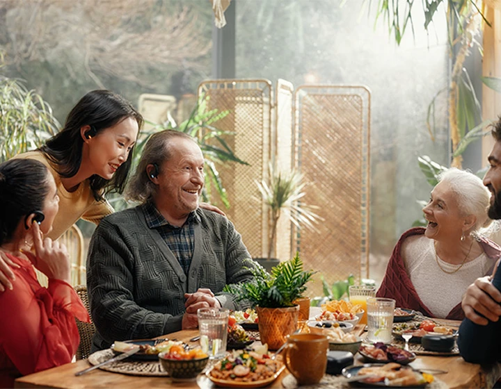 Una familia sentada alrededor de una mesa, con los auriculares E1 puestos, riendo y charlando entre ellos.