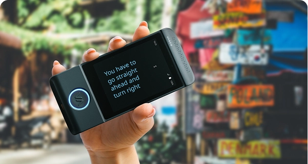 Primer plano de una mano que sostiene en posición horizontal un traductor electrónico Vasco de color negro. La pantalla está en modo de pantalla completa y muestra el texto traducido en una fuente azul grande y nítida: «Tienes que seguir recto y girar a la derecha». El fondo es una escena exterior vibrante y desenfocada en la que se ve un poste de colores cubierto de señales de dirección hacia diversos países, entre ellos Polonia, Chile y Dinamarca. La luz del sol es intensa y resalta el texto del dispositivo para facilitar su lectura. 