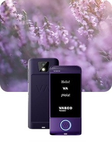 Imagen de presentación del producto en la que se muestra un dispositivo Vasco Translator de color morado desde dos ángulos, sobre un fondo de tela oscura con textura. El dispositivo de la izquierda muestra la vista trasera, en la que destacan la lente de la cámara con flash y el logotipo «VA» en relieve. El dispositivo de la derecha muestra la vista frontal con la pantalla iluminada, en la que aparecen los saludos «Hello!», «VA», «¡Hola!» y el logotipo de «VASCO Translator». Un botón circular situado en la parte inferior del dispositivo está resaltado con una luz azul brillante. La composición se presenta sobre un fondo difuminado y de ensueño de flores de brezo moradas bañadas por una suave luz solar.