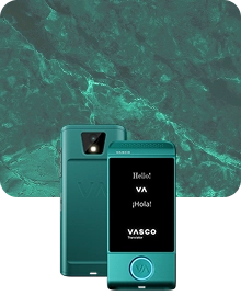 Imagen de presentación del producto en la que se muestra un dispositivo Vasco Translator de color turquesa desde dos ángulos, sobre un fondo de tela oscura con textura. El dispositivo de la izquierda muestra la vista trasera, en la que destacan la lente de la cámara con flash y el logotipo «VA» en relieve. El dispositivo de la derecha muestra la vista frontal con la pantalla iluminada, en la que aparecen los saludos «Hello!», «VA», «¡Hola!» y el logotipo de «VASCO Translator». Un botón circular situado en la parte inferior del dispositivo está resaltado con una luz azul brillante. El fondo presenta un lujoso patrón de mármol verde con vetas de color claro.
