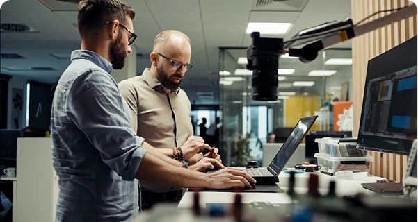 Dos hombres con barba y gafas trabajan juntos en un escritorio alto en una oficina o laboratorio moderno. Uno de ellos, vestido con una camisa azul claro, utiliza un ordenador portátil, mientras que el otro, con una camisa de color beige, está de pie a su lado y señala con un gesto un pequeño componente electrónico que tiene en la mano. El escritorio cuenta con un monitor grande, diversas herramientas electrónicas y una cámara montada en un brazo articulado. Al fondo se ve un espacio de trabajo luminoso y diáfano con mamparas de cristal. 