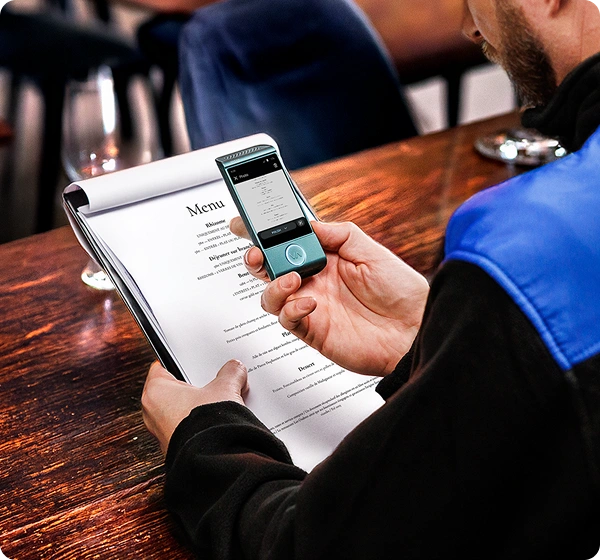 Un primer plano desde un ángulo elevado de una persona sentada en una mesa de madera en un restaurante, utilizando un traductor electrónico portátil para leer el menú. La persona sostiene un menú en papel blanco con la mano izquierda, mientras que con la derecha sujeta un pequeño traductor Vasco de color verde azulado sobre él. La pantalla del dispositivo muestra una versión digital del menú traducido al polaco. La persona lleva una chaqueta negra con un panel azul brillante en el hombro. El fondo está ligeramente desenfocado, mostrando el cálido interior de un restaurante. 