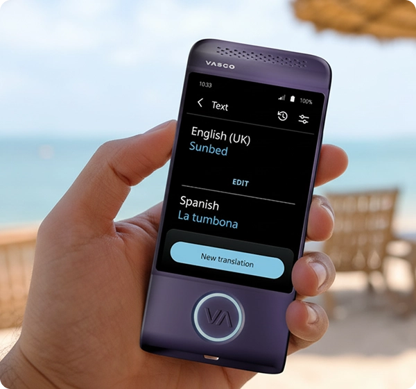 Primer plano de una mano que sostiene un traductor electrónico Vasco de color morado. La pantalla muestra la traducción de un texto del inglés (Reino Unido) al español, en la que la palabra «Sunbed» aparece traducida como «La tumbona». El fondo es una escena de playa luminosa y soleada, con una vista difuminada del océano, arena dorada, varias tumbonas y una sombrilla de paja. 