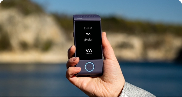 Primer plano de una mano que sostiene un traductor electrónico Vasco de color morado, con un fondo exterior luminoso. En la pantalla aparecen los saludos «Hello!», «¡Hola!» y el logotipo de «VASCO Translator». El fondo muestra una imagen desenfocada de un paisaje costero soleado, con aguas azules y acantilados rocosos de tonos claros bajo un cielo despejado. La persona que sostiene el dispositivo lleva una chaqueta con mangas de textura gris claro o plateada. 