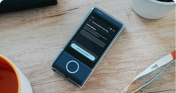 Un primer plano desde un ángulo elevado de un traductor electrónico Vasco de color azul oscuro, dentro de una funda protectora transparente, apoyado sobre una mesa de madera clara. La pantalla del dispositivo está activa y muestra un menú de «Compartir conversación» con un campo para introducir una dirección de correo electrónico y un botón que dice «Compartir conversación». Alrededor del dispositivo, sobre la mesa, se ven el borde de una taza de café naranja, una maceta blanca y un cordón blanco con la palabra «Hej!» impresa. La escena está muy bien iluminada, lo que crea una estética limpia y moderna. 