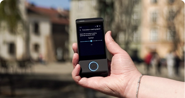 Primer plano de la mano de una persona que sostiene un traductor electrónico Vasco de color negro, con un fondo difuminado de una plaza urbana con edificios históricos. La pantalla del dispositivo muestra los ajustes de «Velocidad de lectura de la traducción», con un control deslizante situado en «Normal» y una nota que dice: «Ajuste la velocidad de lectura de la traducción moviendo el control deslizante». La persona que sostiene el dispositivo lleva una pulsera plateada. La luz brillante del día resalta la interfaz de la pantalla, lo que facilita la visualización de los ajustes. 