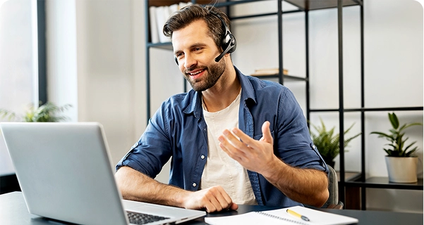 Un hombre de aspecto afable, con barba y cabello oscuro, está sentado ante un escritorio y lleva unos auriculares profesionales con micrófono. Sonríe y gesticula con una mano mientras mira la pantalla de un ordenador portátil; parece estar en medio de una videollamada o prestando asistencia técnica. Viste una camisa azul abotonada sobre una camiseta blanca. Sobre el escritorio que tiene delante hay un ordenador portátil, un cuaderno y un bolígrafo amarillo. El fondo es un espacio de oficina luminoso y moderno, con una estantería minimalista de metal negro y plantas en macetas verdes. 