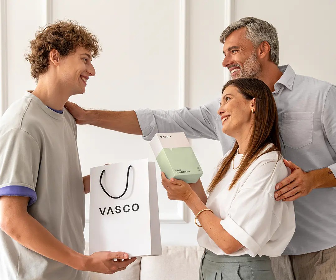 Una escena conmovedora en la que tres personas sonríen juntas. A la izquierda, un joven de pelo rizado sostiene una bolsa de la compra blanca de «VASCO». En el centro, una mujer sostiene una caja blanca y verde claro con la etiqueta «Vasco Translator M4» y mira feliz al joven. Detrás de ella, un hombre con el pelo canoso sonríe cálidamente, con una mano sobre el hombro de la mujer y la otra sobre el hombro del joven. Se encuentran en una habitación luminosa con paredes de color neutro y un sofá al fondo. 