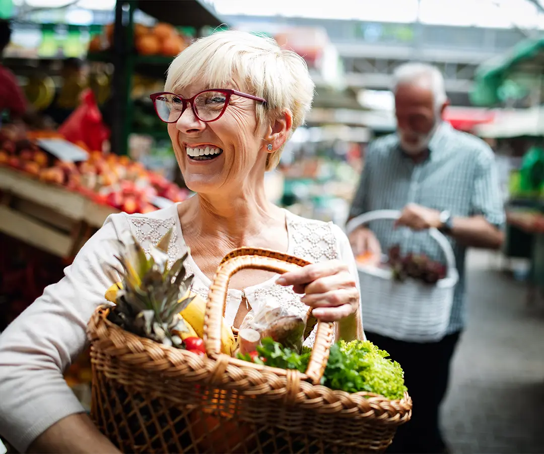 Una mujer mayor sonriente, con el pelo rubio corto y gafas rojas, lleva una cesta de mimbre llena de fruta y verdura fresca en un mercado al aire libre. En el fondo, difuminado, se ve a otra persona comprando entre los puestos de productos frescos.