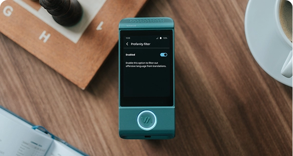 Una toma desde arriba de un traductor electrónico Vasco de color verde azulado apoyado sobre una mesa de madera. La pantalla del dispositivo está encendida y muestra el menú de configuración del «Filtro de lenguaje soez», con el interruptor en la posición «Activado». Debajo del interruptor, se lee la siguiente descripción: «Activa esta opción para filtrar el lenguaje ofensivo de las traducciones». El dispositivo está rodeado de objetos cotidianos, como un tablero de ajedrez de madera con una pieza oscura, la esquina de un cuaderno abierto y una taza de café blanca, todo ello capturado bajo una luz suave y natural. 