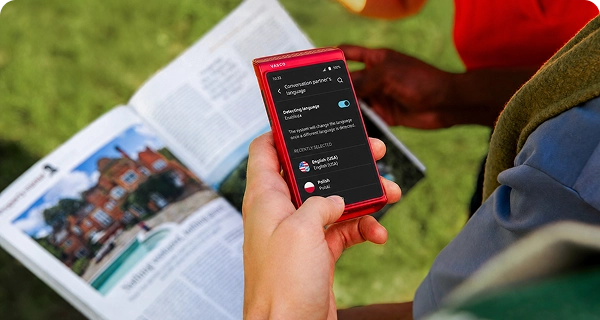 Una persona utiliza un Traductor Q1 rojo para hacer una foto de un periódico. En la pantalla aparece el menú de selección de idioma para traducir el texto detectado automáticamente del periódico.